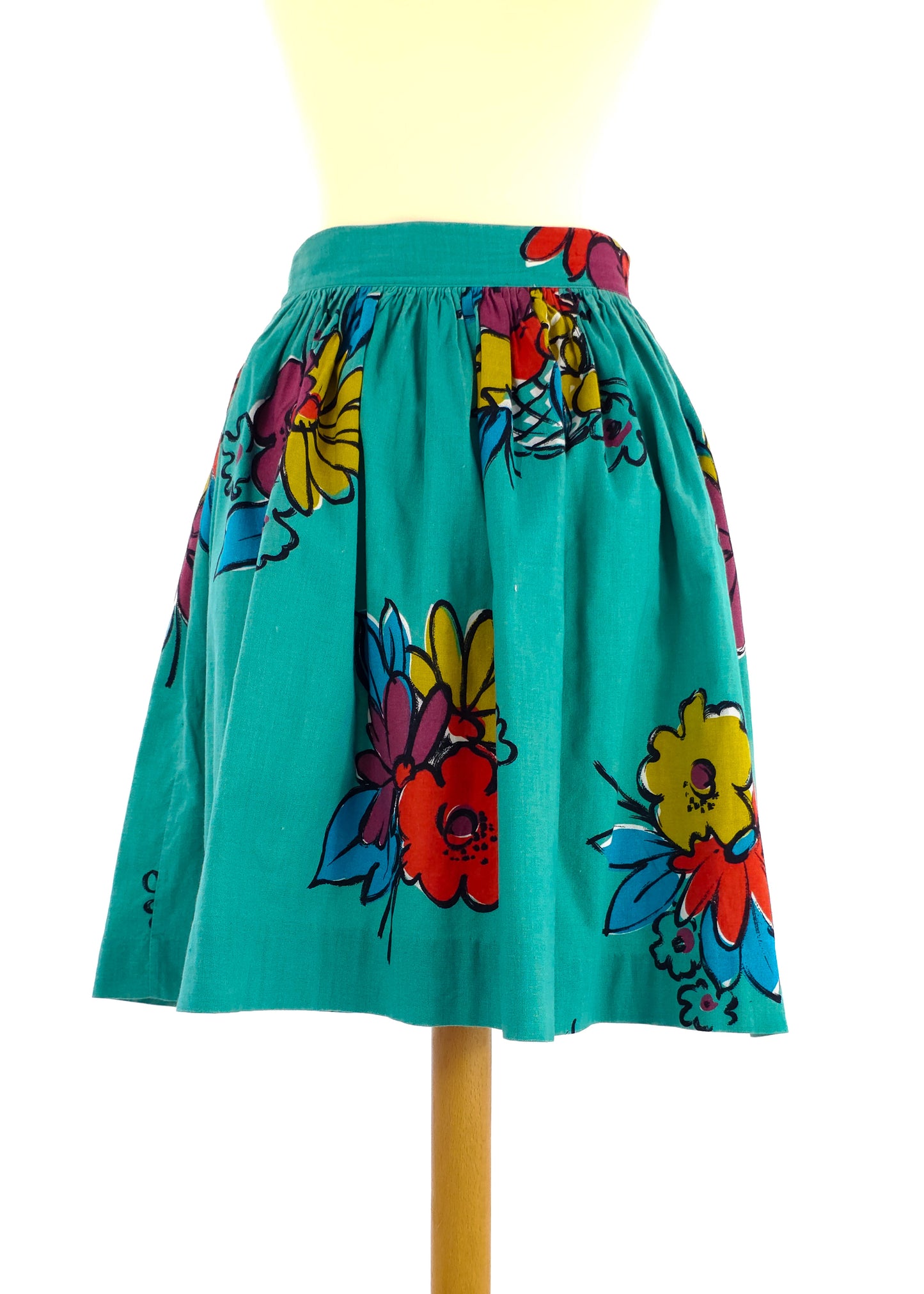 A teal skirt with a high waist and vibrant floral print in red, yellow, and blue, displayed on a mannequin. The skirt is knee-length and has a gathered waistband.
