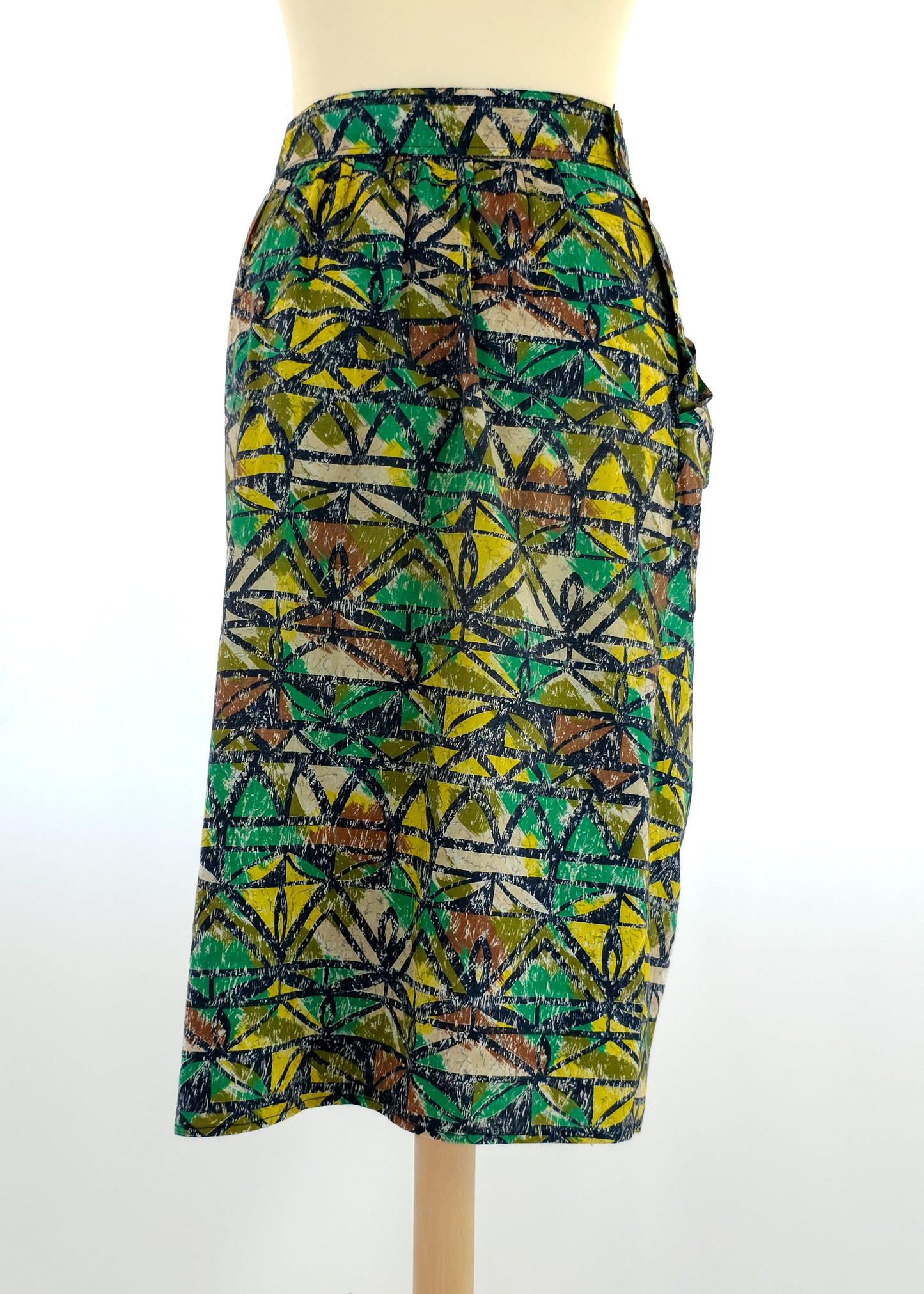 A knee-length vintage skirt with a vibrant green, yellow, and brown geometric pattern displayed on a mannequin. The skirt has a fitted waist and slight pleats.