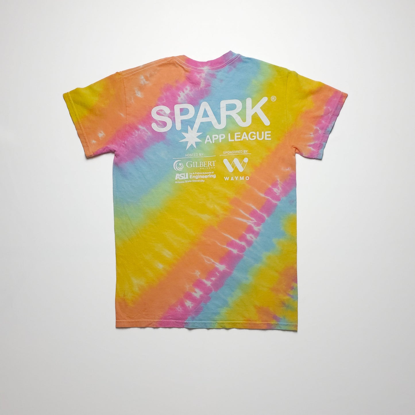 A colorful tie-dye t-shirt with a white "SPARK App League" logo on the back. The shirt features diagonal swirls of orange, blue, yellow, and pink.