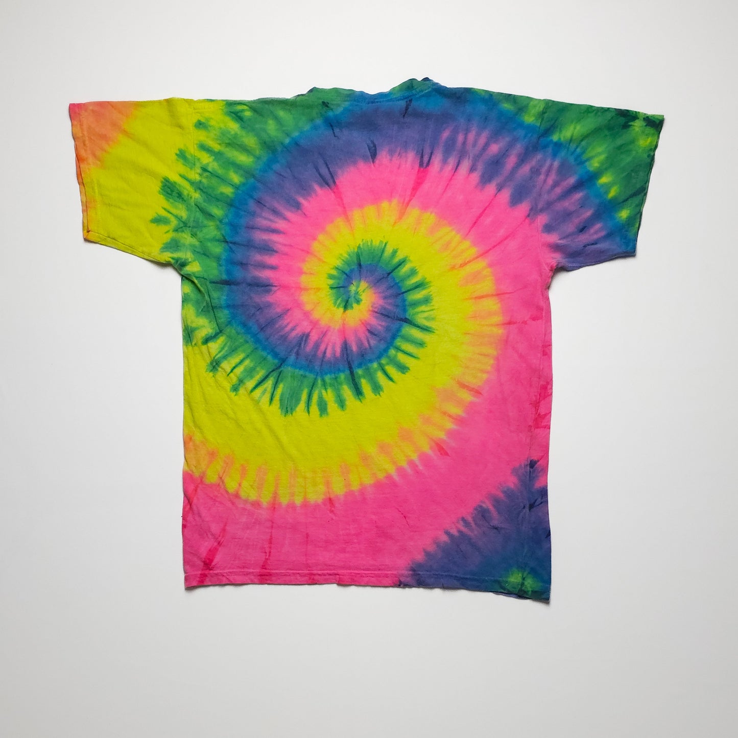 A vibrant tie-dye T-shirt featuring a spiral pattern in pink, yellow, green, and blue. The shirt is laid flat against a plain white background.