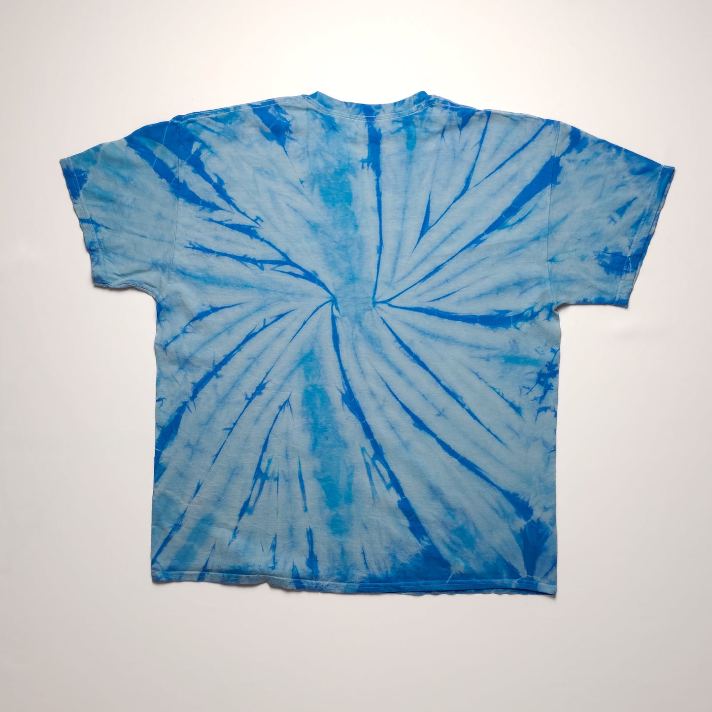 A blue tie-dye T-shirt with a spiral pattern on a plain white background, showing the back side. The shirt features various shades of blue.