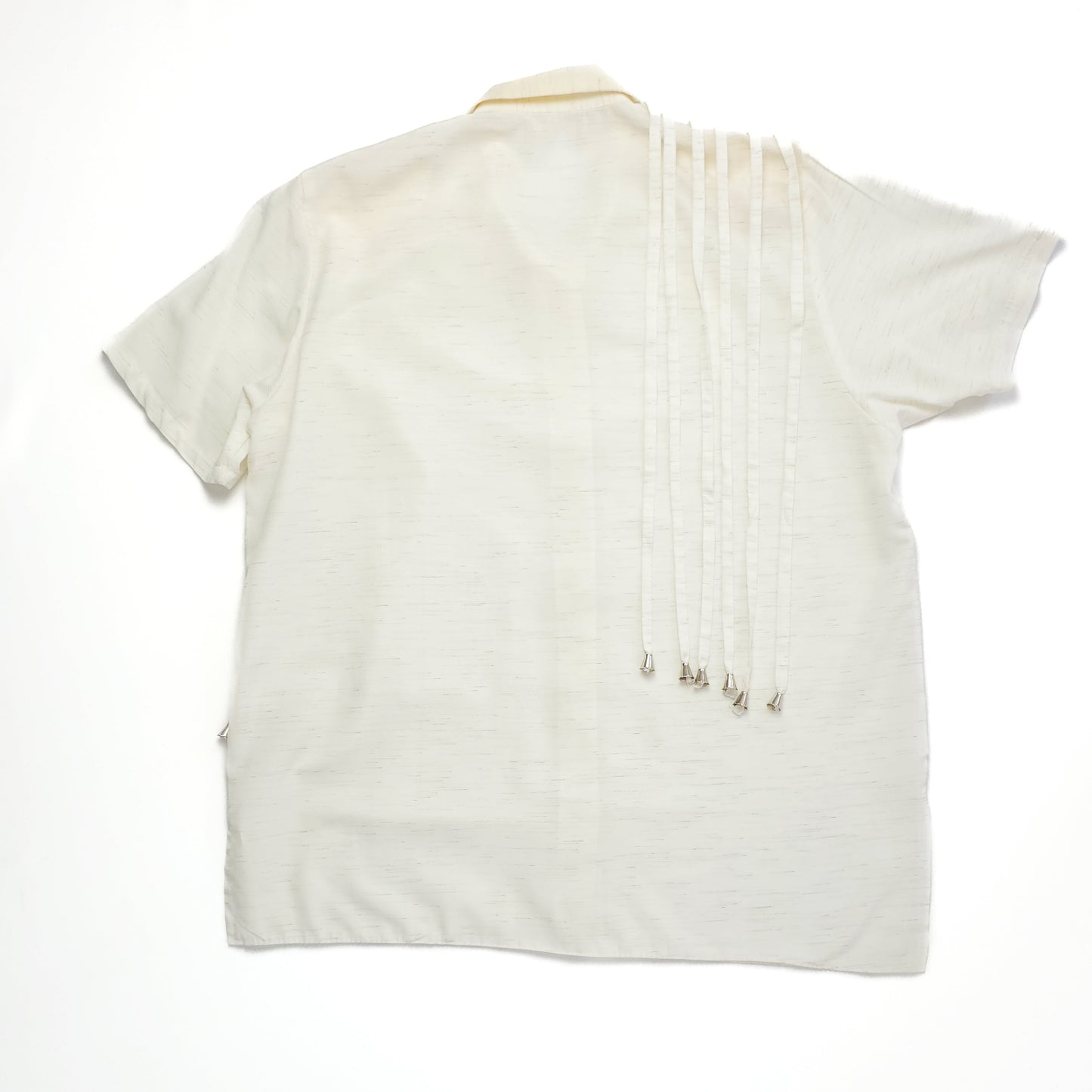 A white short-sleeve shirt with fringe detailing on the right shoulder, laid flat on a plain surface. The fabric appears lightweight and slightly textured.