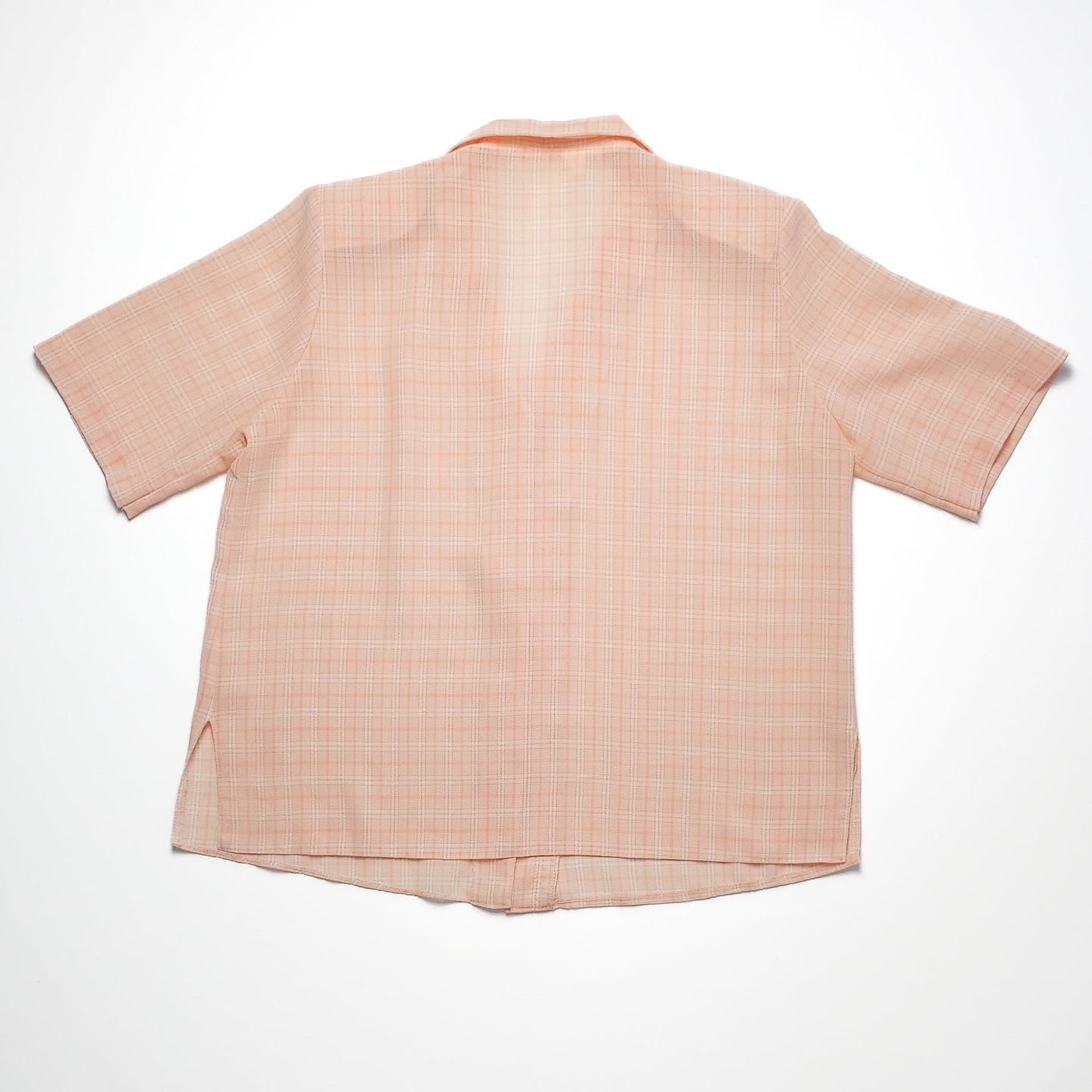 A light peach short-sleeve button-up shirt with a subtle checkered pattern. The fabric is lightweight, and the hem has small side slits for comfort.