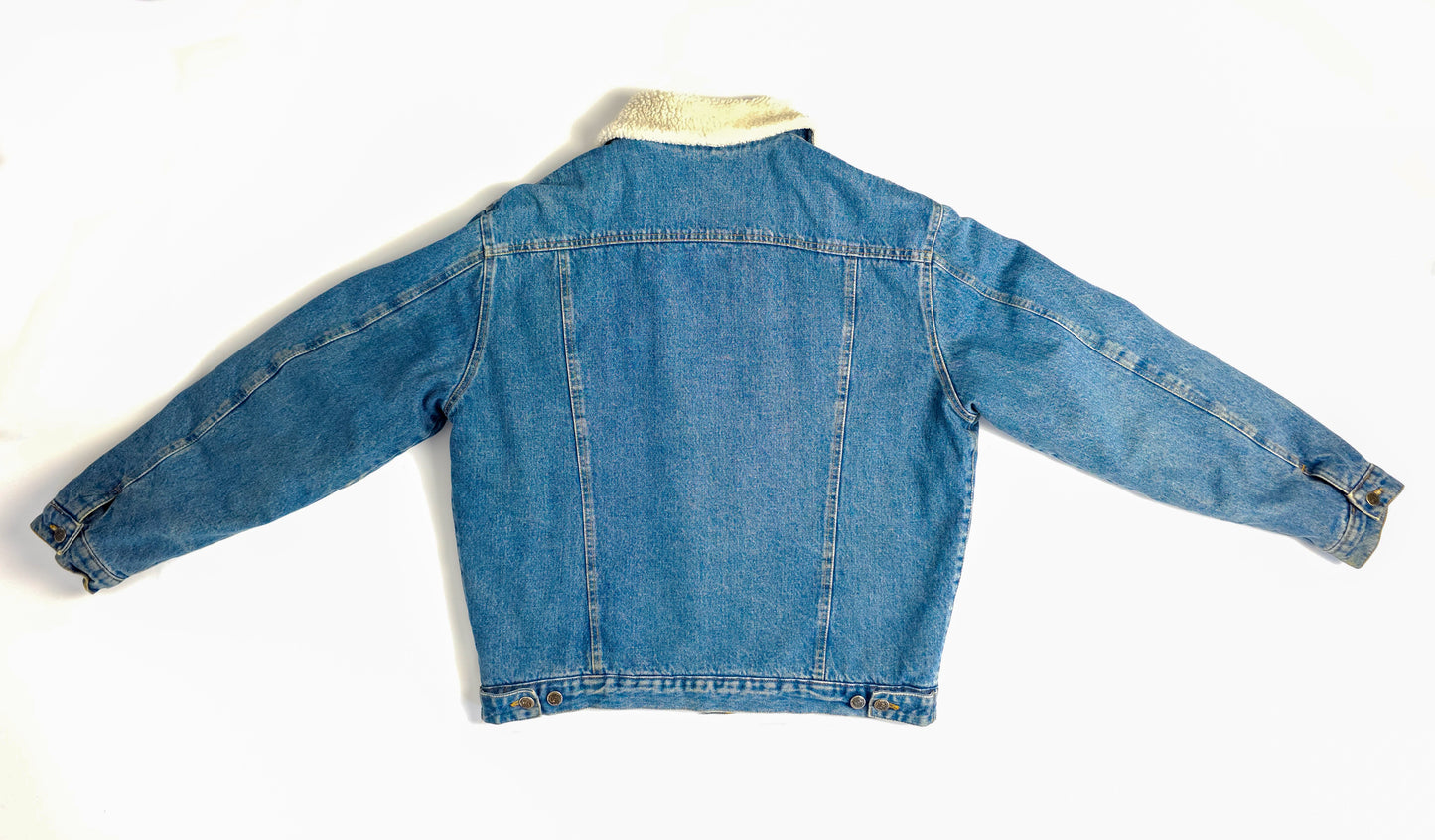 A back view of a blue denim jacket with long sleeves and a light-colored shearling collar. The jacket has a straight silhouette and button details.