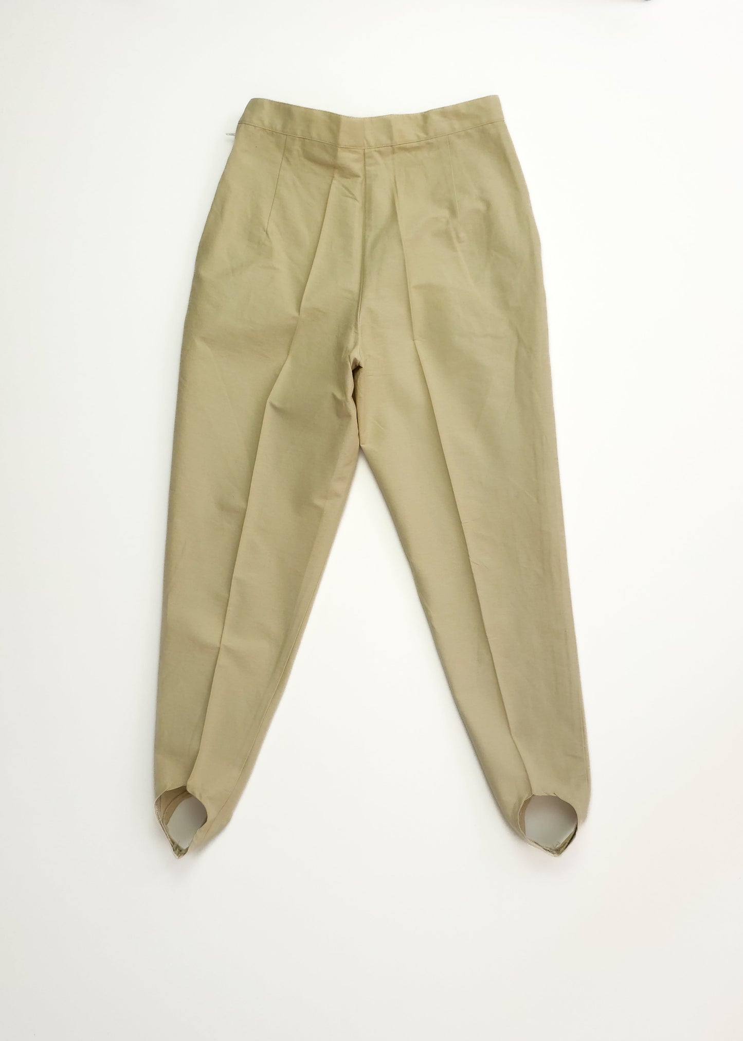 Beige high-waisted stirrup pants with pleats and tapered legs, designed for a snug fit. The pants are laid flat against a white background.