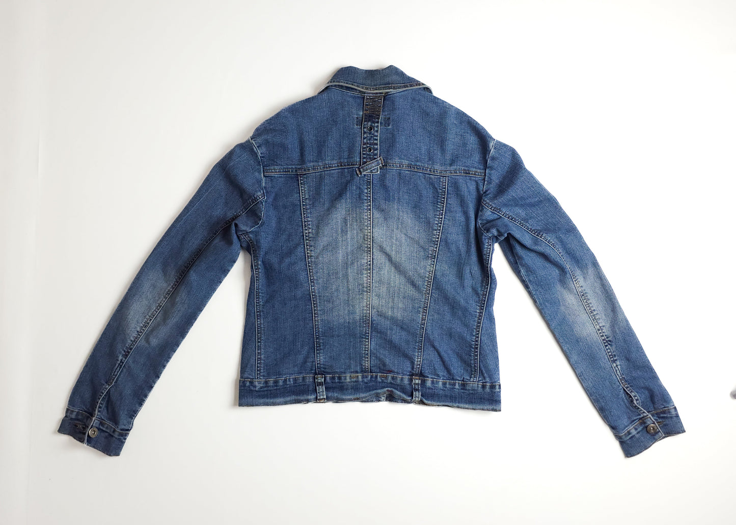 A blue denim jacket with long sleeves, laid flat facing down. The jacket has a classic collar, buttoned cuffs, faded details, and visible seam stitching.