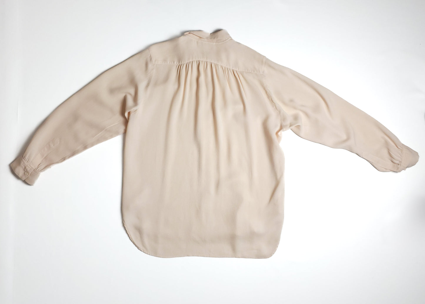 A light beige, long-sleeved women's blouse with a loose fit and a collared neckline, spread flat on a white surface. The back features subtle pleating.