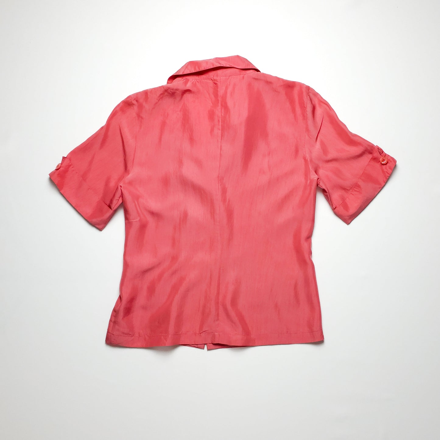 A coral pink short-sleeve blouse with a collared neckline, buttoned cuffs, and a smooth back design, laid flat on a white surface.