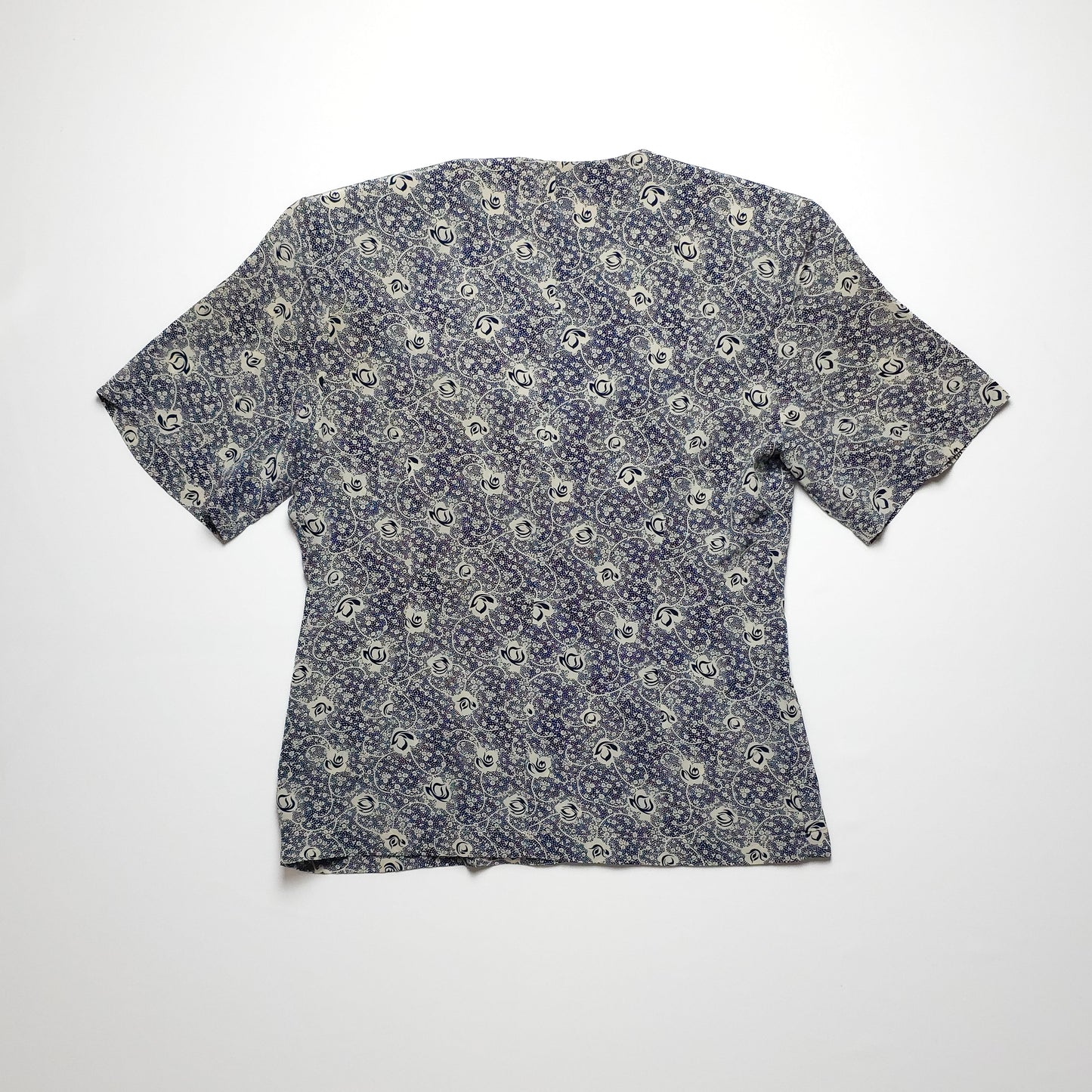 A short-sleeved, vintage floral-patterned blouse in shades of blue and white, laid flat on a plain white background. The fabric has a delicate, intricate design.