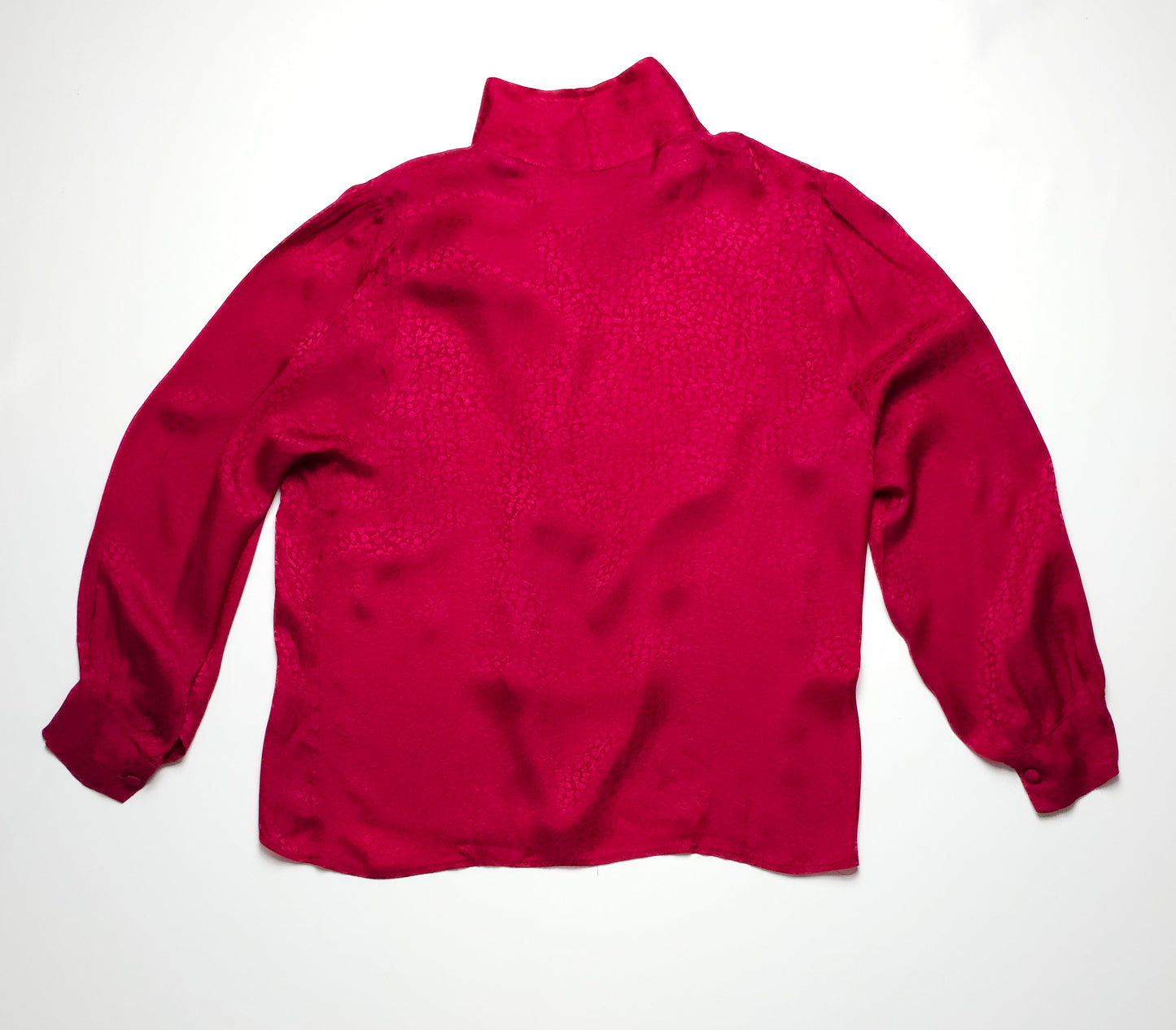 A bright red, long-sleeved blouse with a high collar and buttoned cuffs, laid flat on a white surface. The fabric has a slightly silky texture.