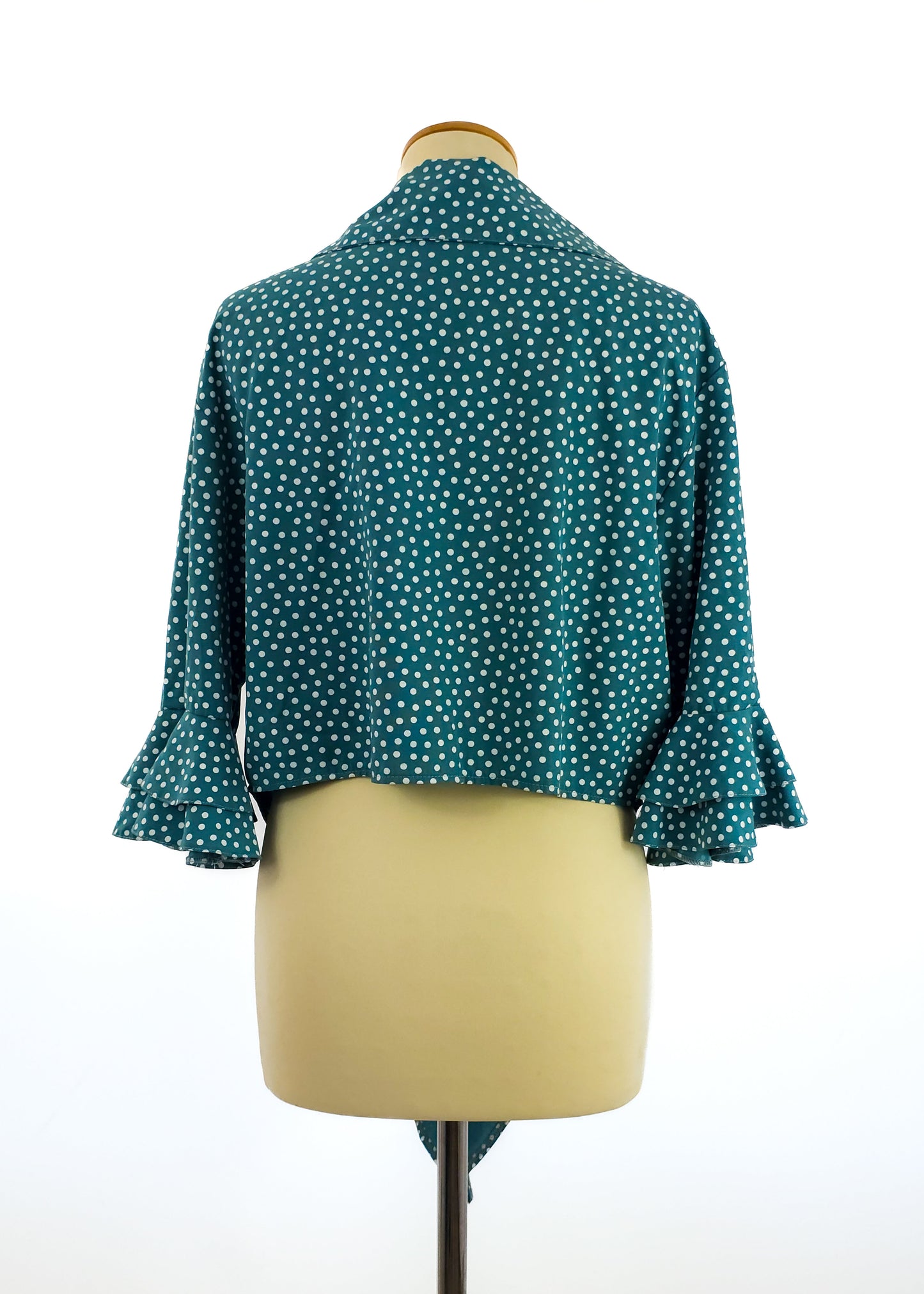 A teal polka dot cropped jacket with ruffled sleeves displayed on a mannequin, viewed from the back. The fabric shows a pattern of small white dots.