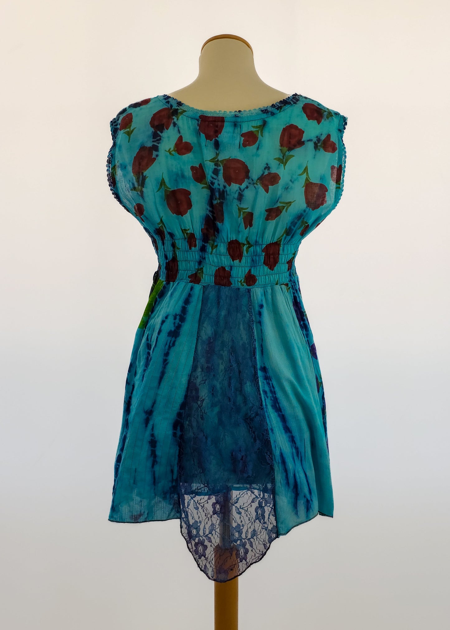 A sleeveless blue dress displayed on a mannequin, featuring a floral pattern on the top, an elastic waist, and lace detailing on the back, with a high-low hem.