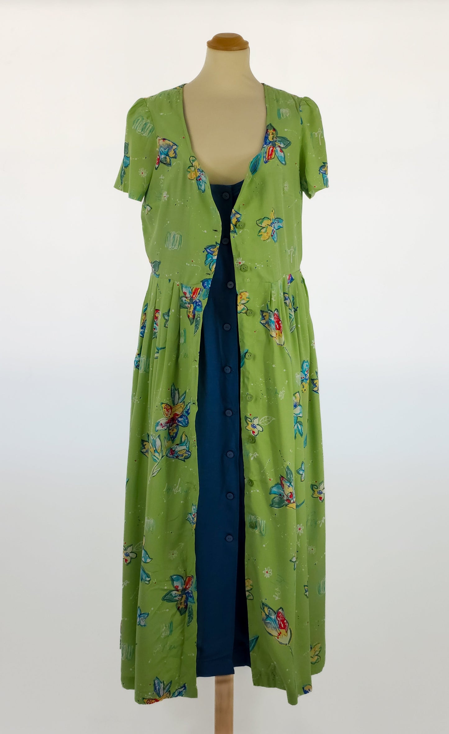 A green floral short-sleeve maxi dress with a button-down front, displayed over a dark blue dress on a mannequin against a white background.