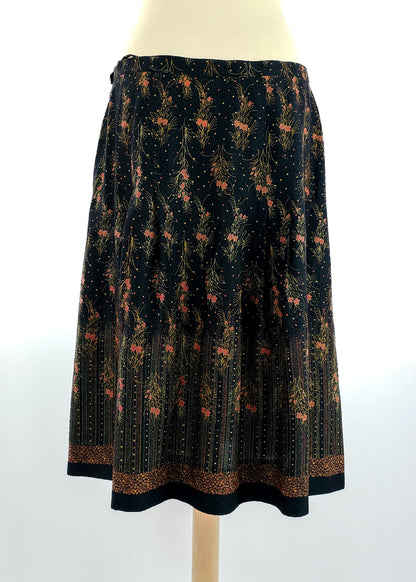 A black floral A-line skirt with a vintage pattern featuring small orange flowers and green leaves. The skirt has a decorative hem and falls below the knee.