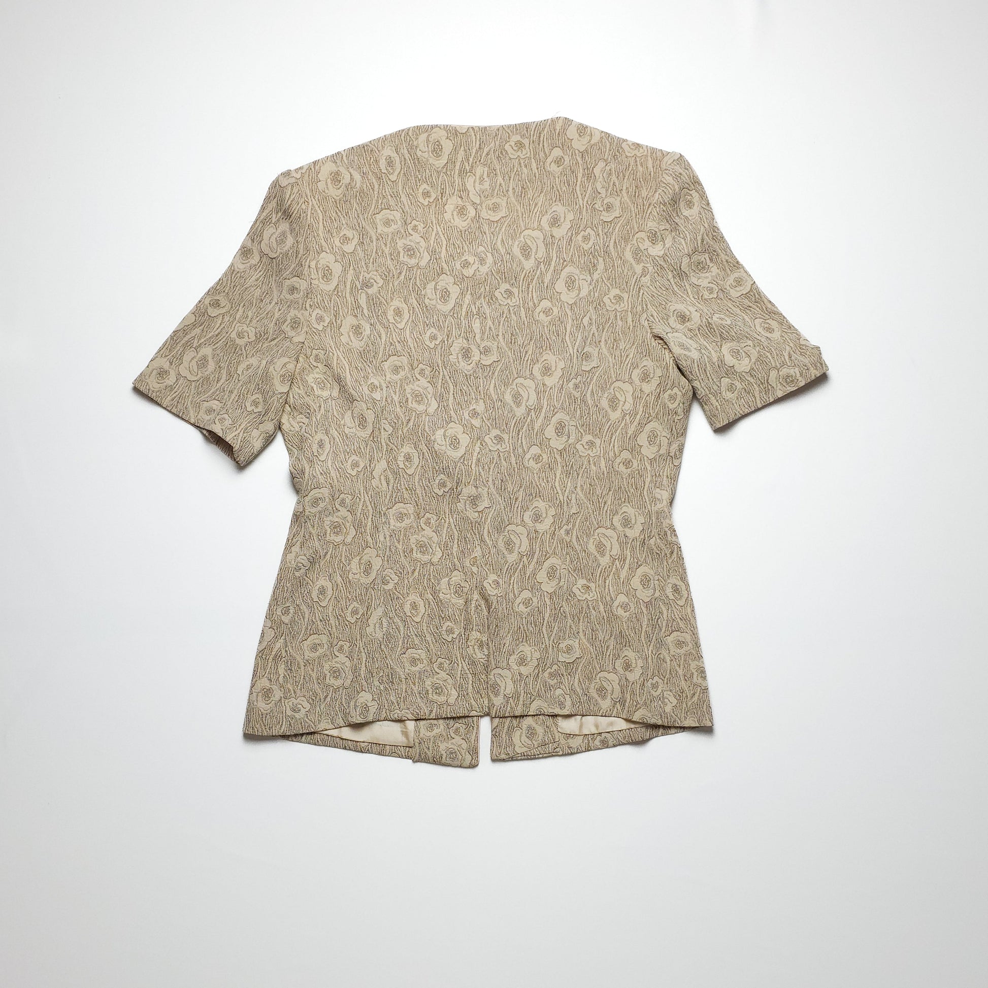 A short-sleeved, textured beige women's blouse with a floral pattern and a rounded neckline, displayed on a white background. The blouse has a gently flared hem.